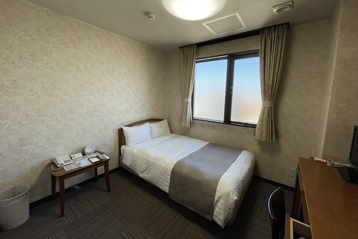 Une chambre d’hôtel avec un lit simple, une table de chevet avec un téléphone et une horloge, une fenêtre avec des rideaux et un bureau avec un ordinateur.