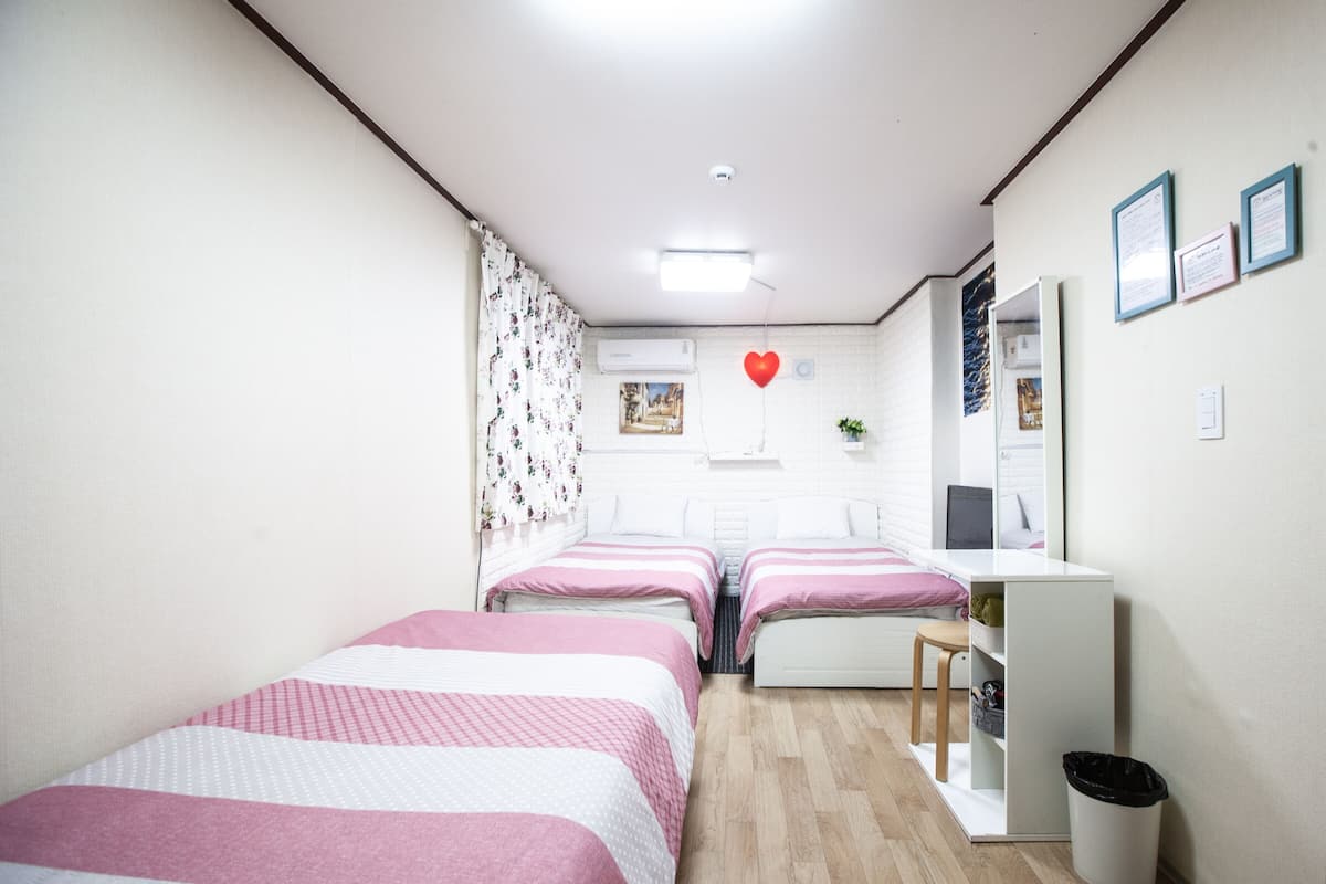 A bedroom with two beds, a desk, a chair, a trash bin, and a wall-mounted heater.