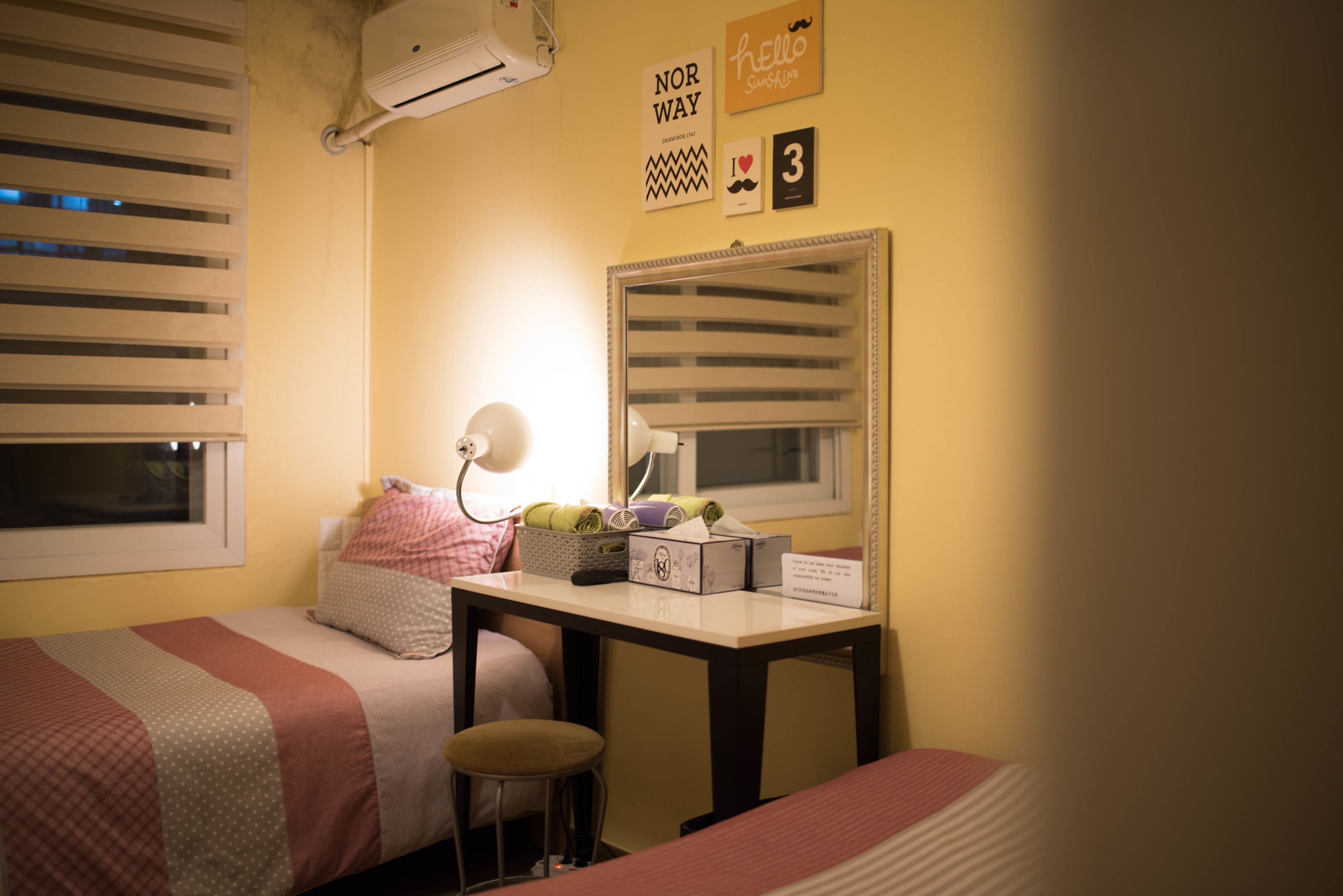 A bedroom with two beds, a desk with a mirror, a lamp, and a window with blinds.