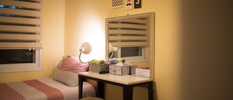A bedroom with two beds, a desk with a mirror, a lamp, and a window with blinds.