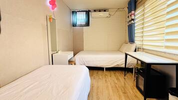 A small, compact room with two single beds, a bedside table, a mirror, a small stool, a window with blinds, and a wall-mounted air conditioner.