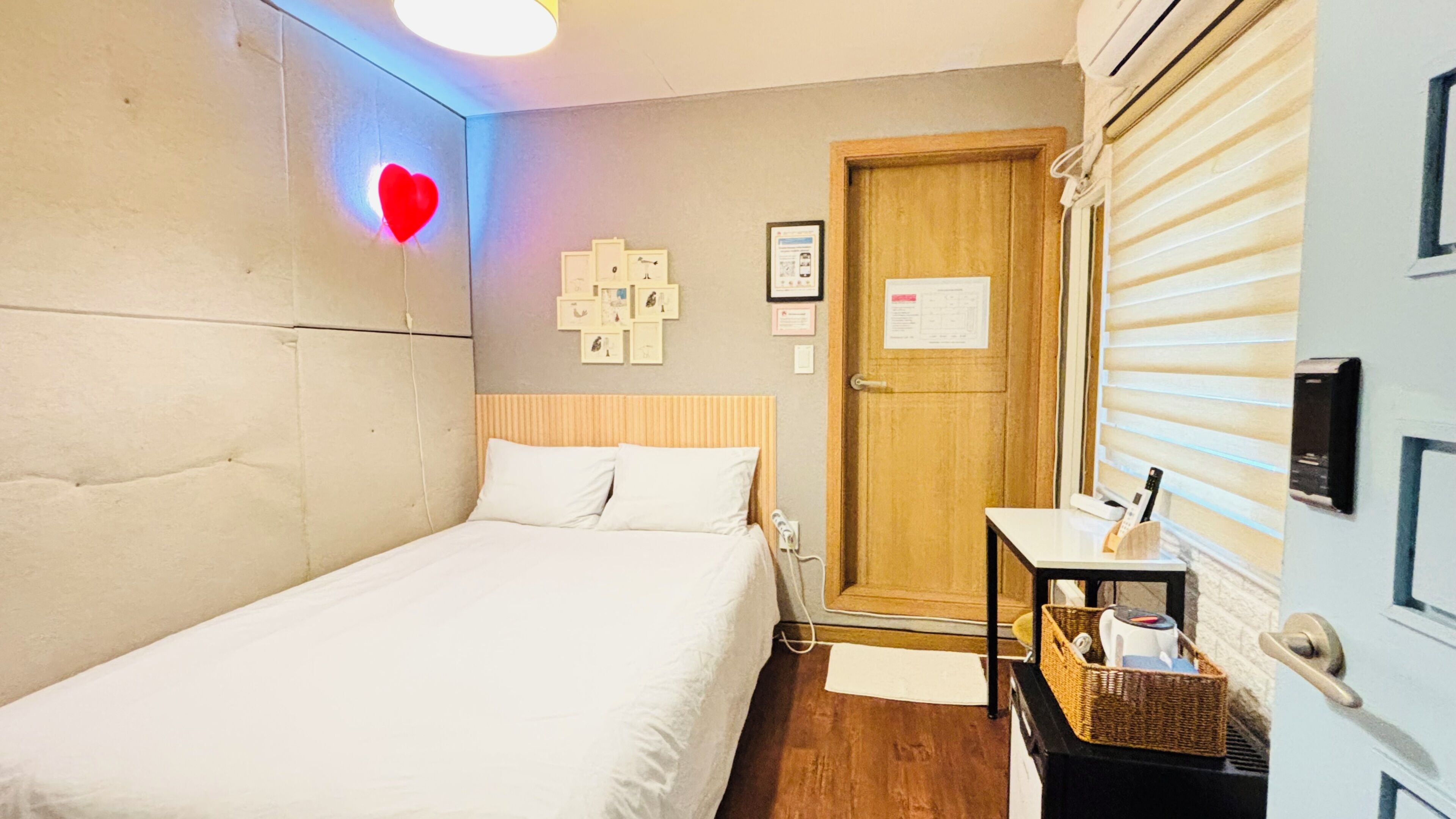 A bedroom with a bed, a bedside table with a lamp, a wall-mounted heater, and a door with a window.