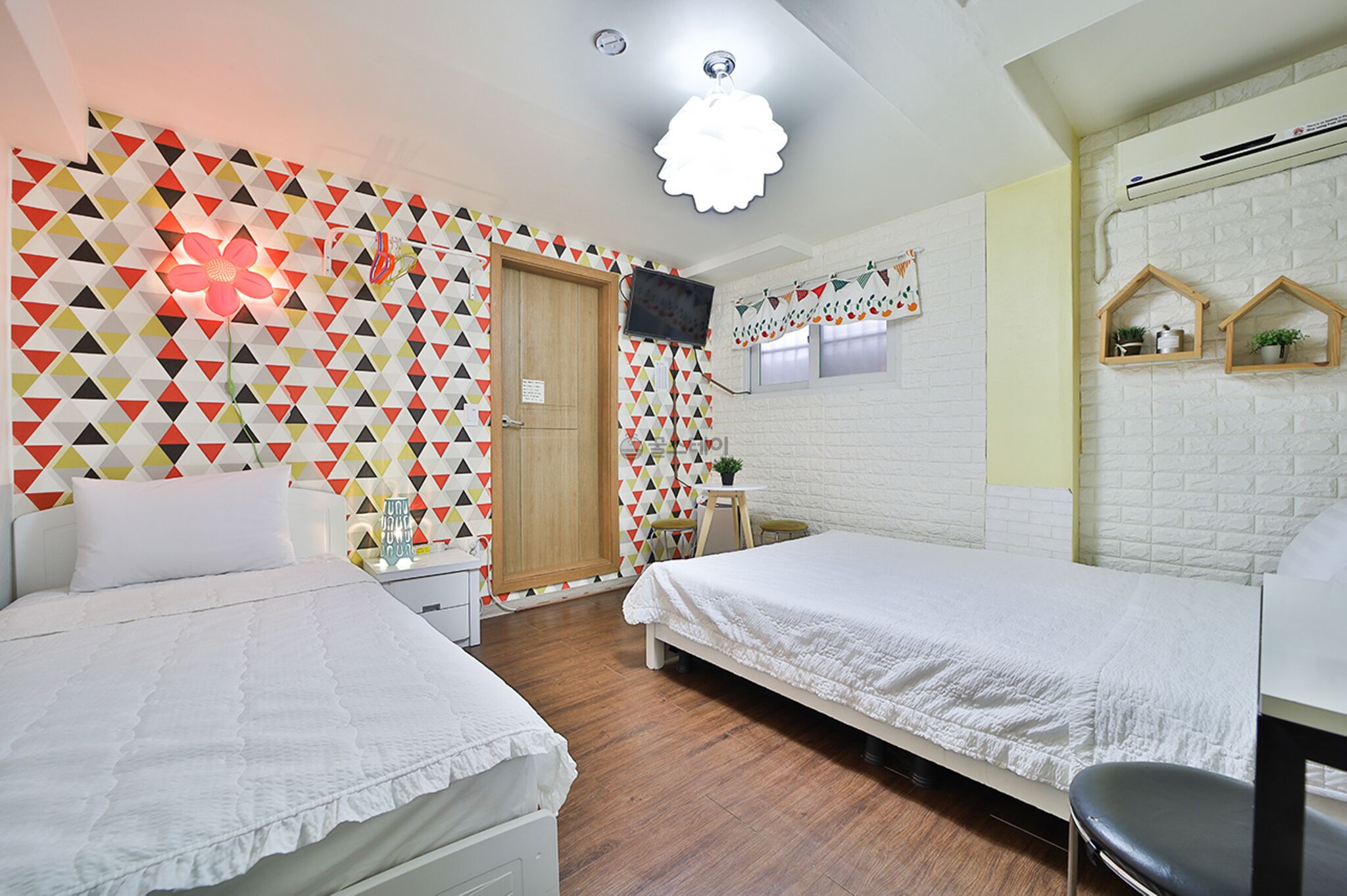 A room with two beds, a wooden door, a TV on the wall, and a small table with a plant.