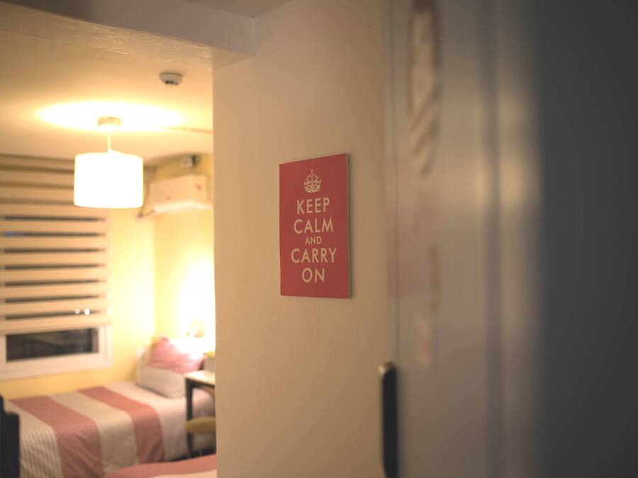 Une chambre avec un lit, une fenêtre avec des stores, une table de chevet et un panneau fixé au mur sur lequel on peut lire : « GARDONS NOTRE CALME ET CONTINUONS ».