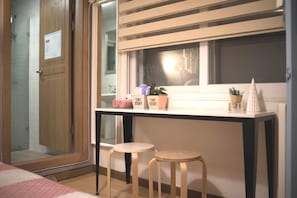 A small room with a wooden door, a window with blinds, a table with two stools, and a bed with a pink and white striped bedspread.