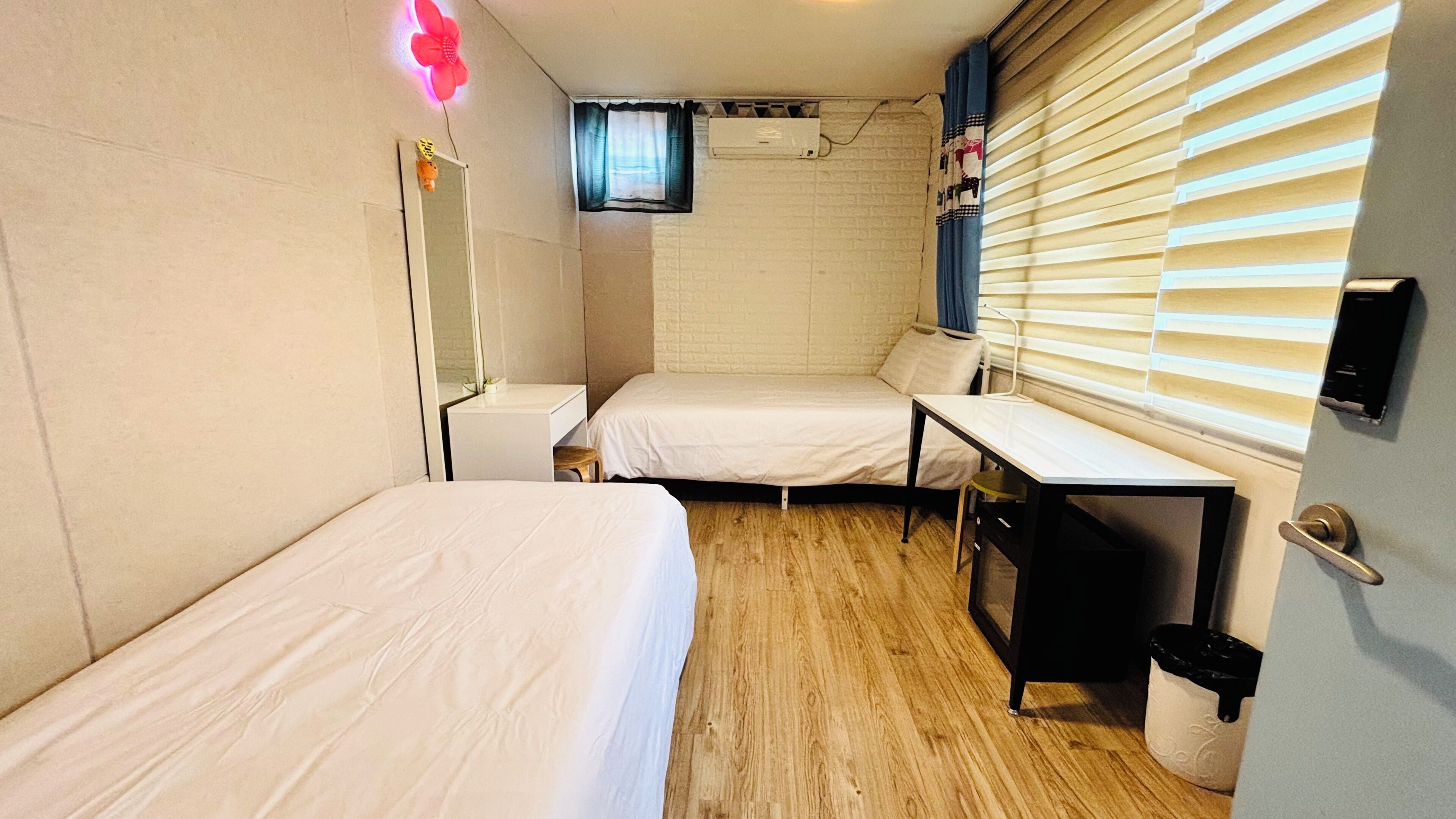 A compact room with two single beds, a small table, a mirror, and a window with blinds.