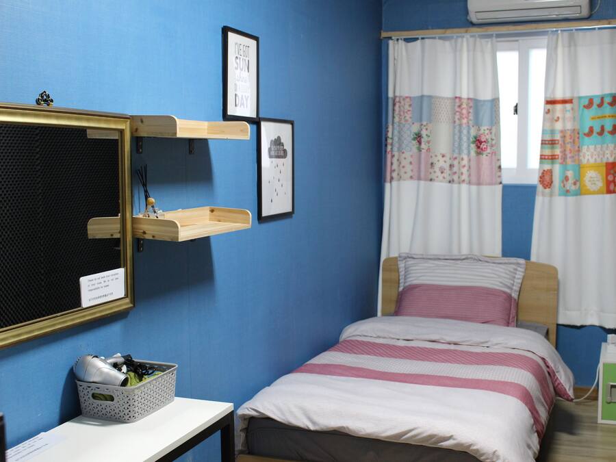Ein Schlafzimmer mit Bett, Nachttisch, Lampe, einem Fenster mit Vorhängen, einem Wandregal und einem gerahmten Bild.