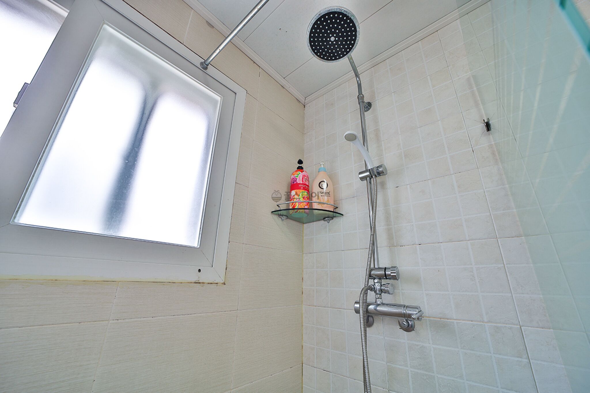 Eine Dusche mit Handbrause, Ablagefläche aus Glas für Toilettenartikel und ein Fenster über der Dusche.