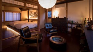A traditional Japanese-style room with tatami flooring, a low wooden ceiling, a bed with white bedding, a black leather chair, a wooden coffee table, and a hanging paper lantern. - Kiraku Kyoto Higashiyama (Kyoto)