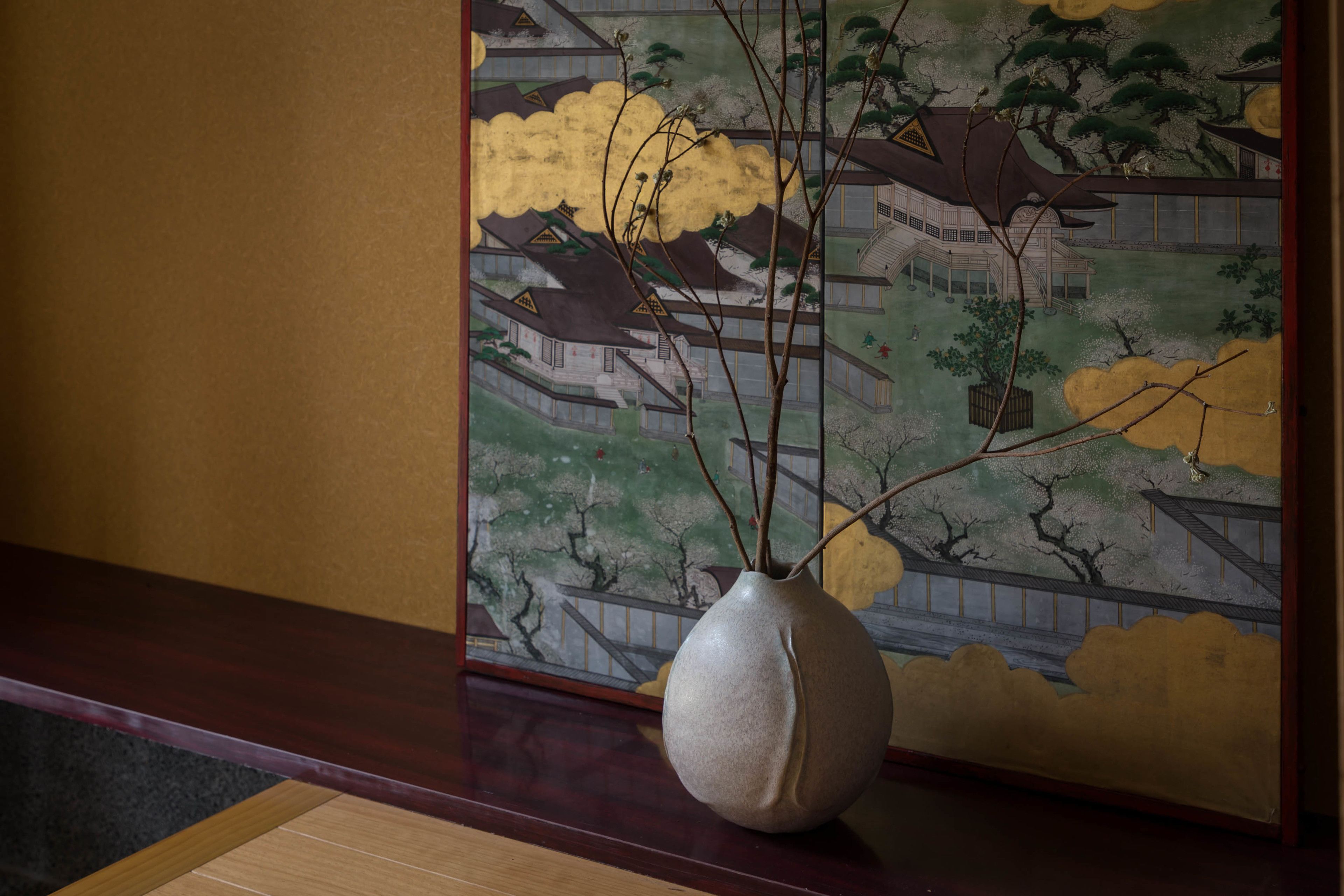 A vase with branches on a wooden table in front of a traditional Japanese painting.