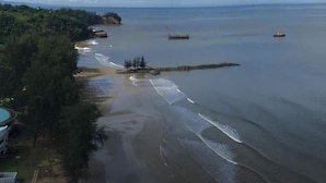 Vista costera con playa arenosa, mar tranquilo y una costa distante con vegetación y edificios.