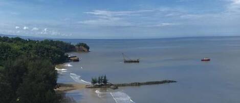 A coastal view with a sandy beach, calm sea, and a distant shoreline with greenery and buildings.