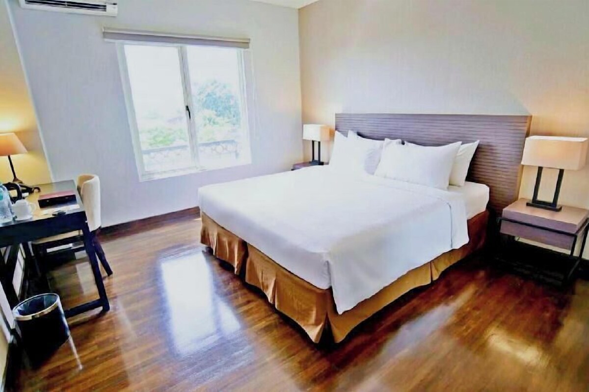 A hotel room with a large bed, two bedside tables with lamps, a desk with a chair, and a window with a view of greenery.