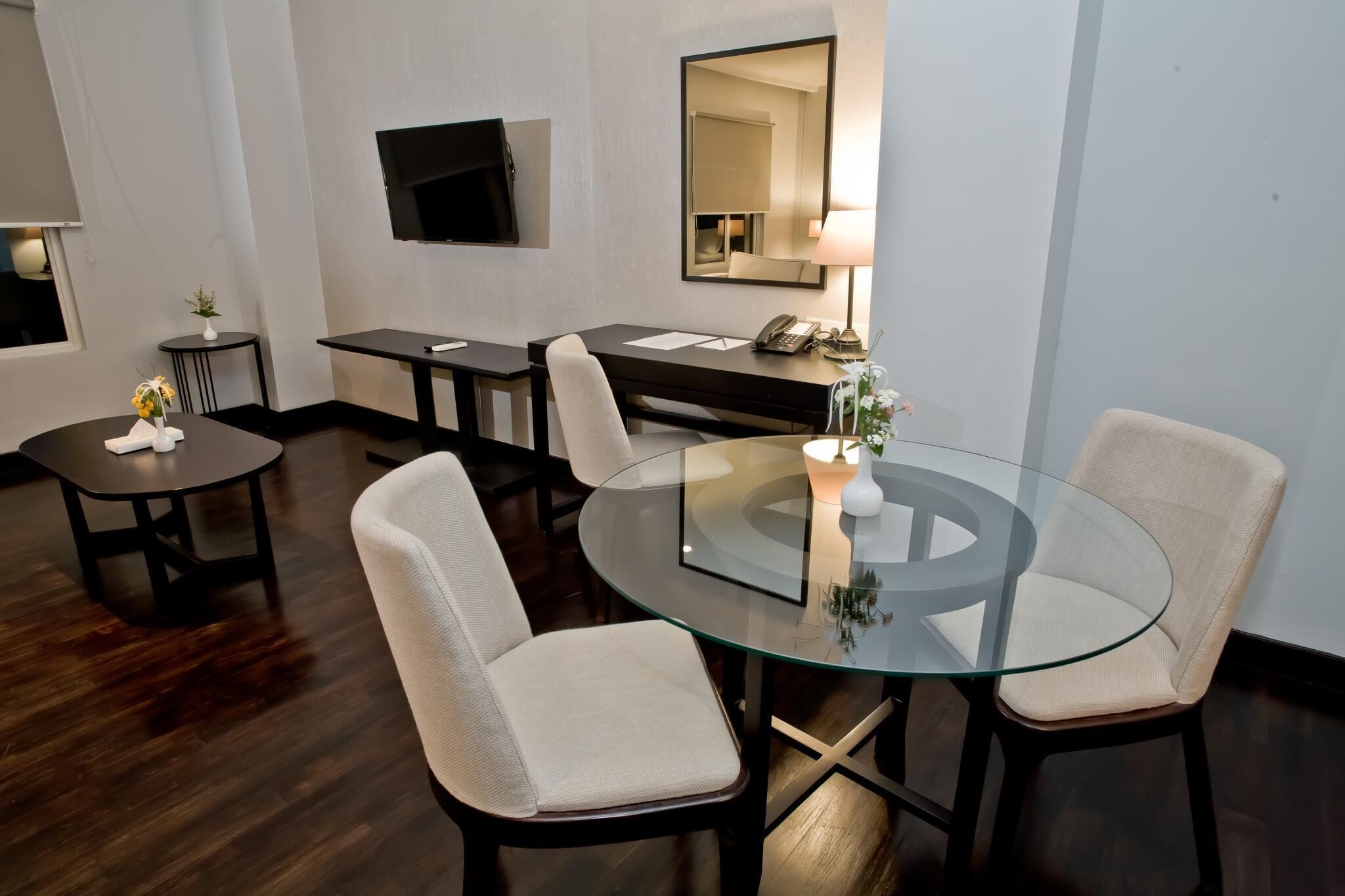 A modern hotel room with a glass dining table, two chairs, a small side table with a vase, a desk with a telephone, and a flat-screen TV mounted on the wall.