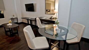 A modern hotel room with a glass dining table, two chairs, a small side table with a vase, a desk with a telephone, and a flat-screen TV mounted on the wall.