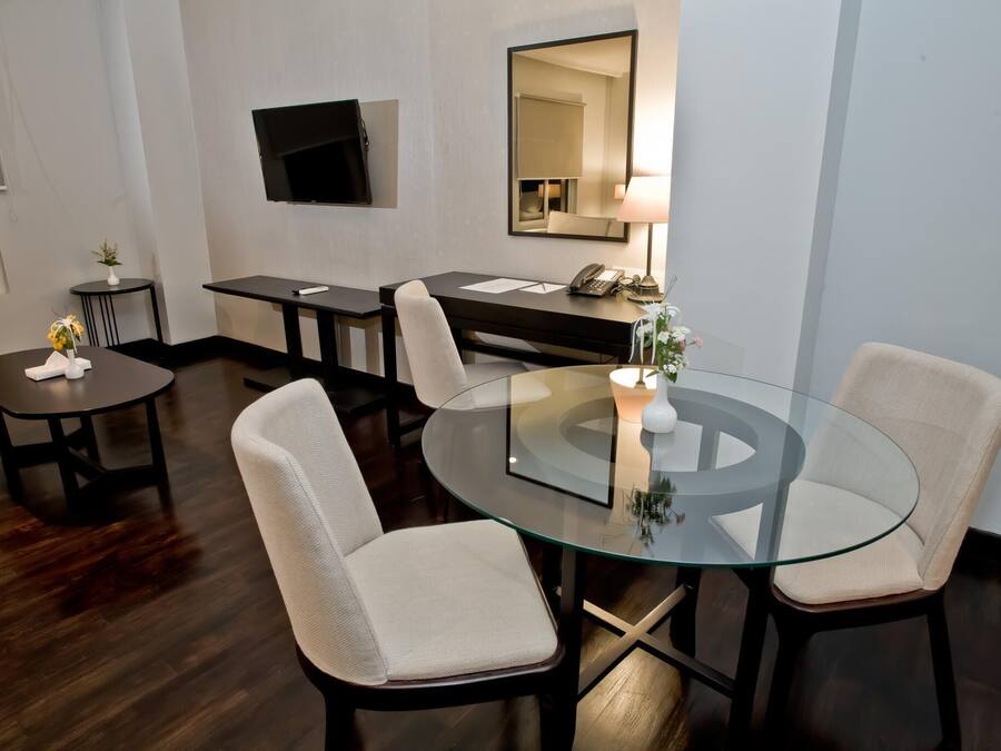 A modern hotel room with a glass dining table, two chairs, a small side table with a vase, a desk with a telephone, and a flat-screen TV mounted on the wall.