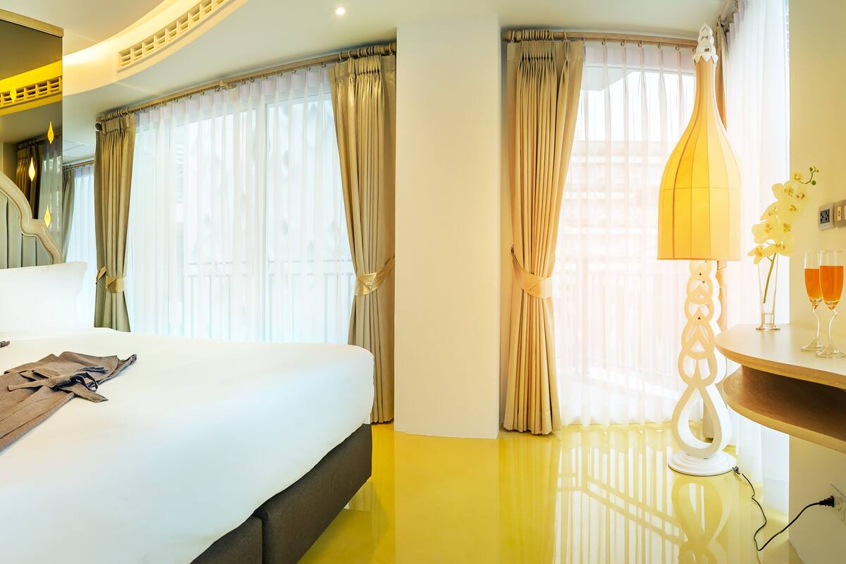 A hotel room with a large bed, a bedside table with a lamp, and a view of the outside through the window.