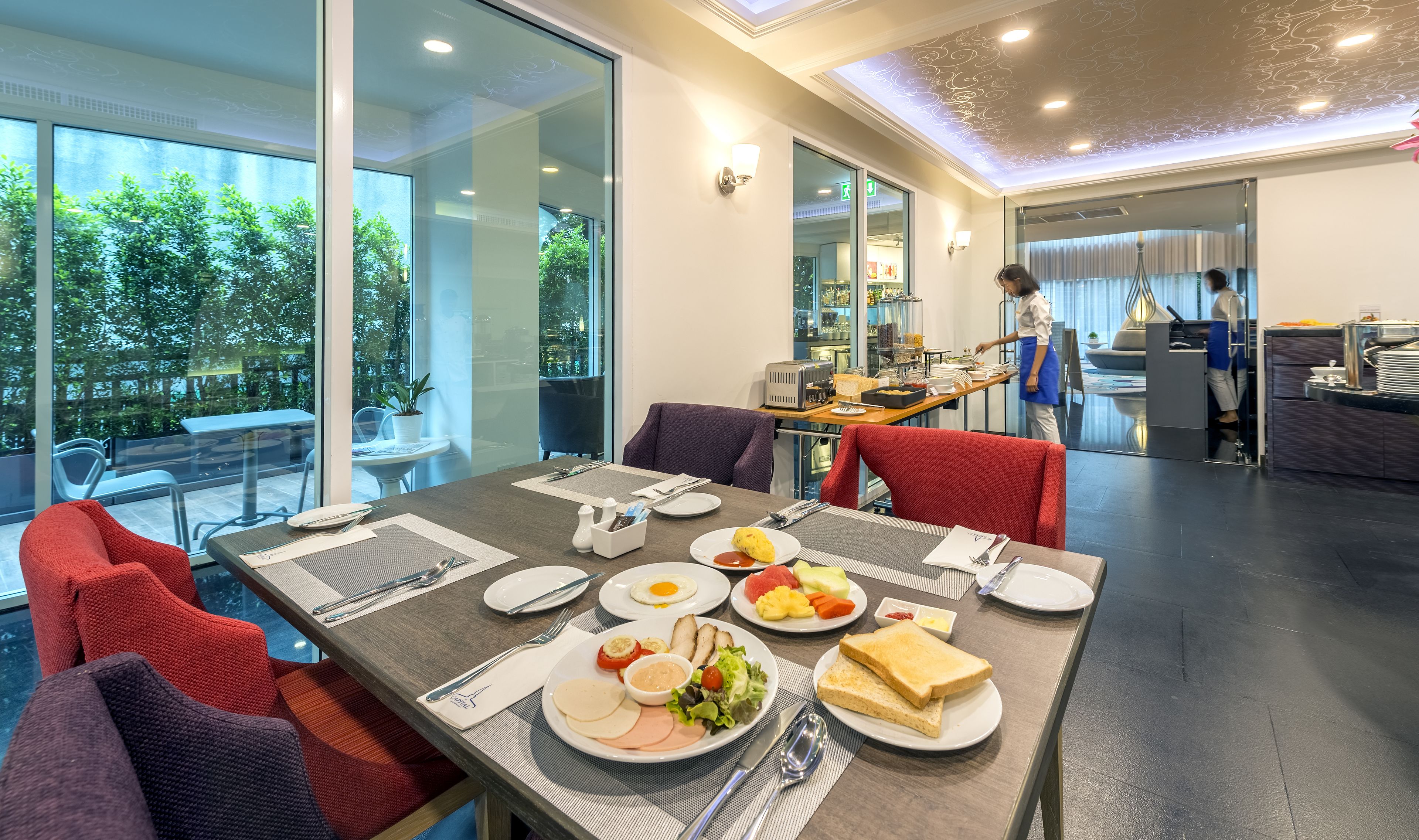 A modern dining area with a large table set for a meal, a kitchen in the background, and a view of greenery outside.