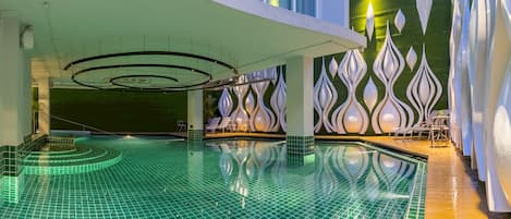 A modern indoor swimming pool with a green tiled floor, white decorative wall panels, and a covered seating area with lounge chairs.