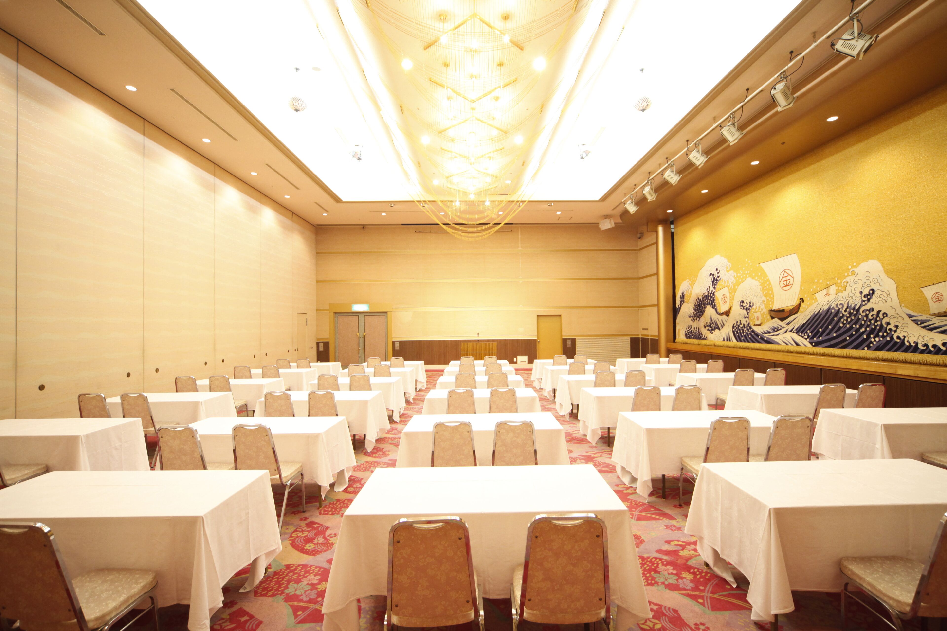 A large conference room with rows of tables and chairs, a high ceiling with chandeliers, and a large mural on the wall.