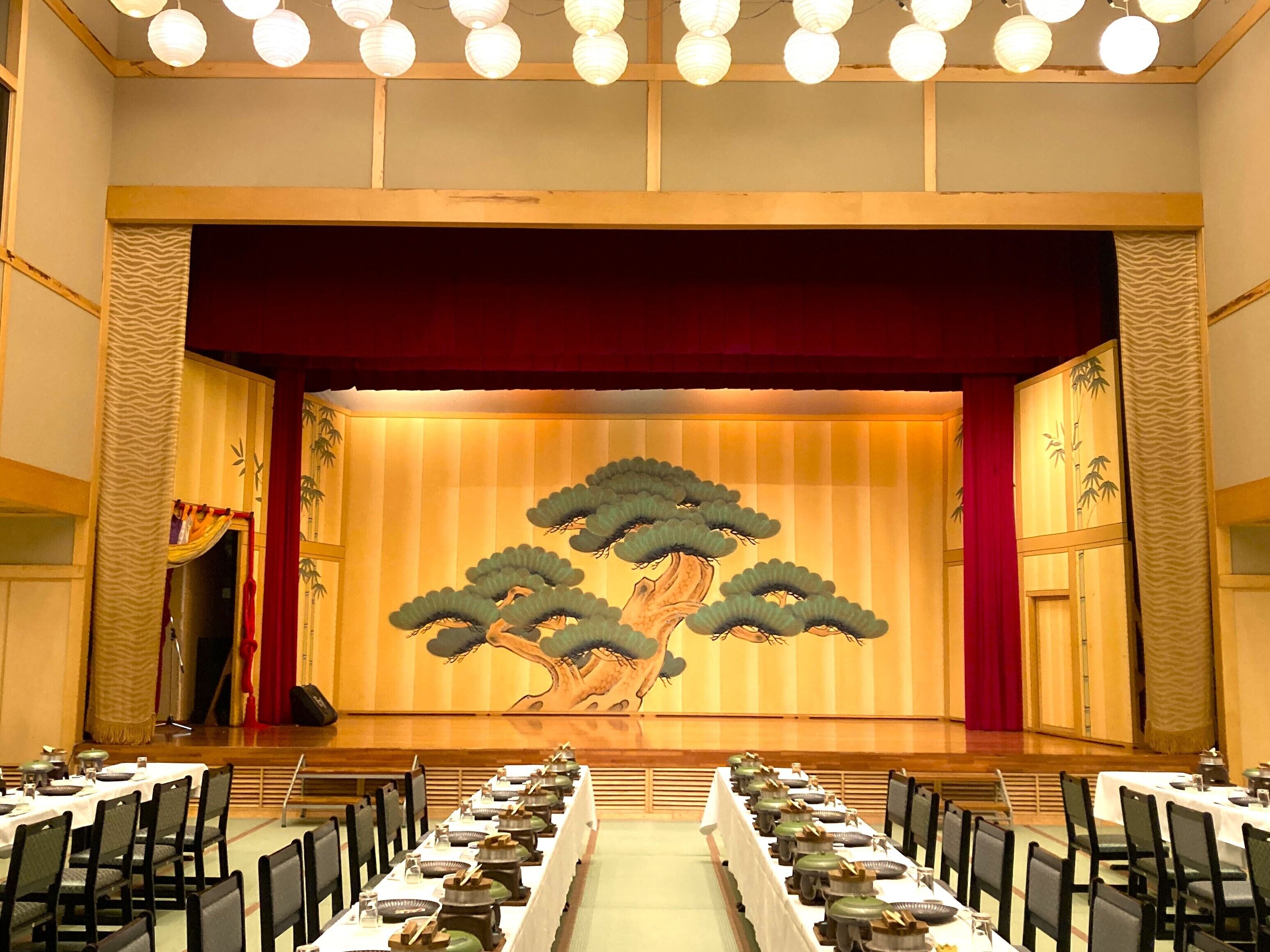 A conference room with a stage featuring a traditional Japanese tree mural, tables set for a meeting, and a red curtain backdrop.