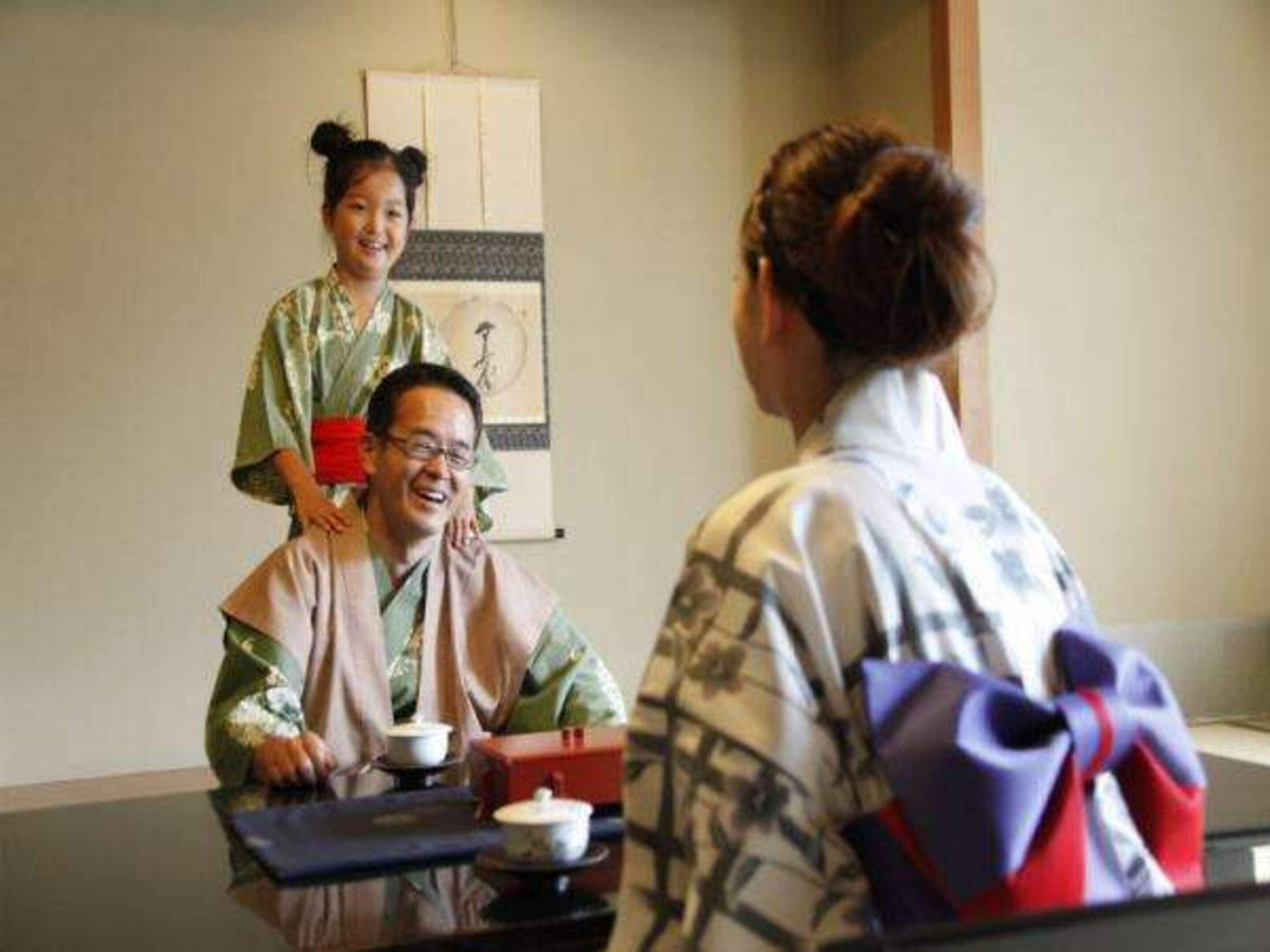 A traditional Japanese room with tatami flooring, a low table with a tea set, and a person in a kimono.
