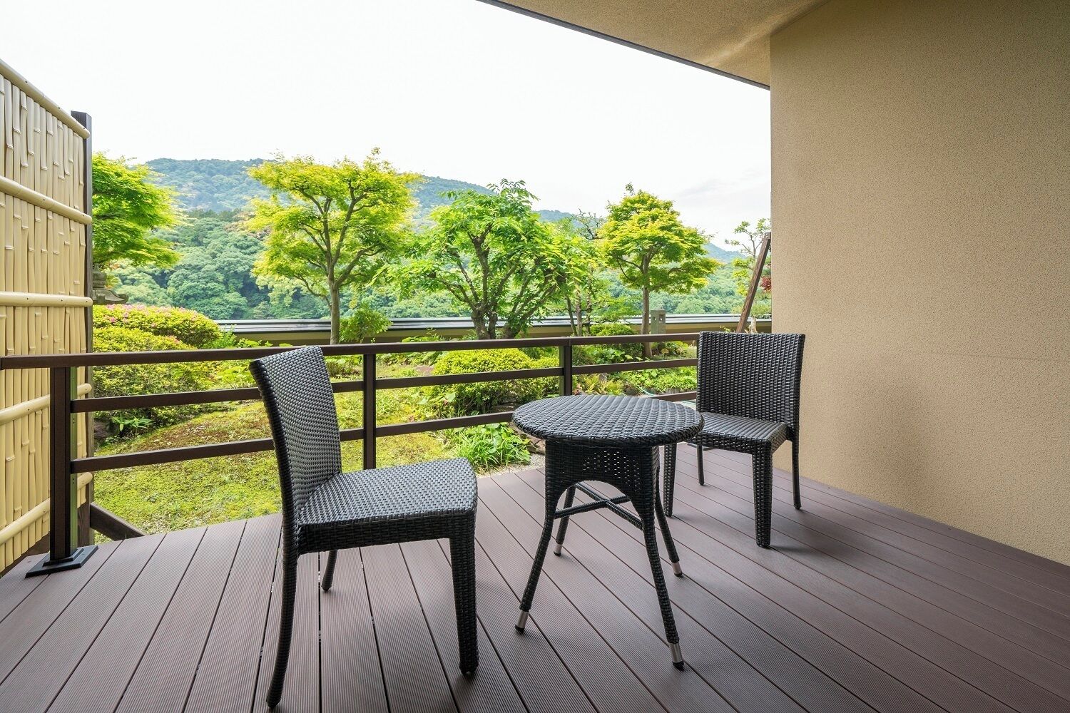 A balcony with two wicker chairs and a small round table, overlooking a lush green landscape with trees and hills.