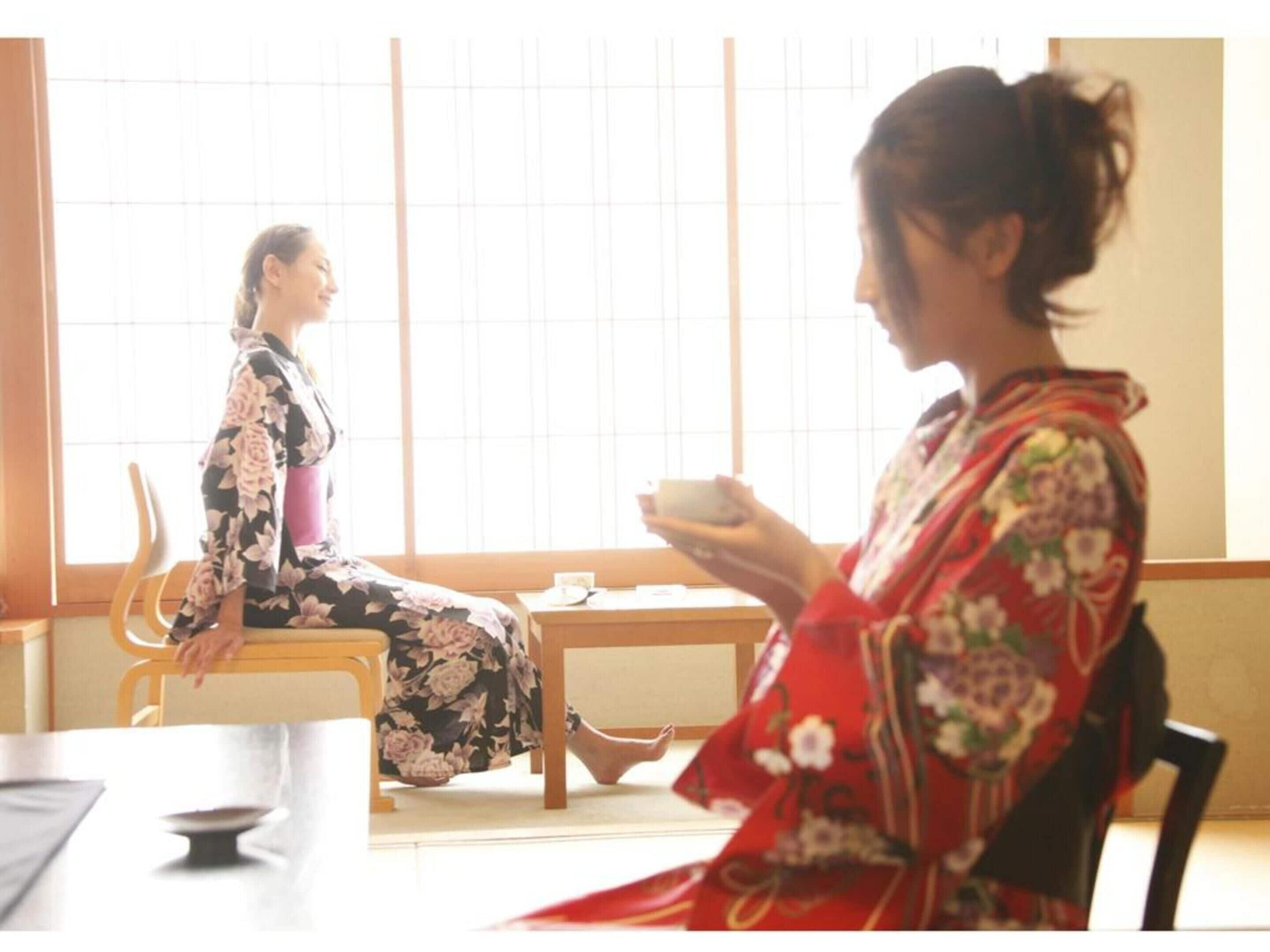 Two people in traditional attire sitting across from each other at a table in a room with large windows and a low table.