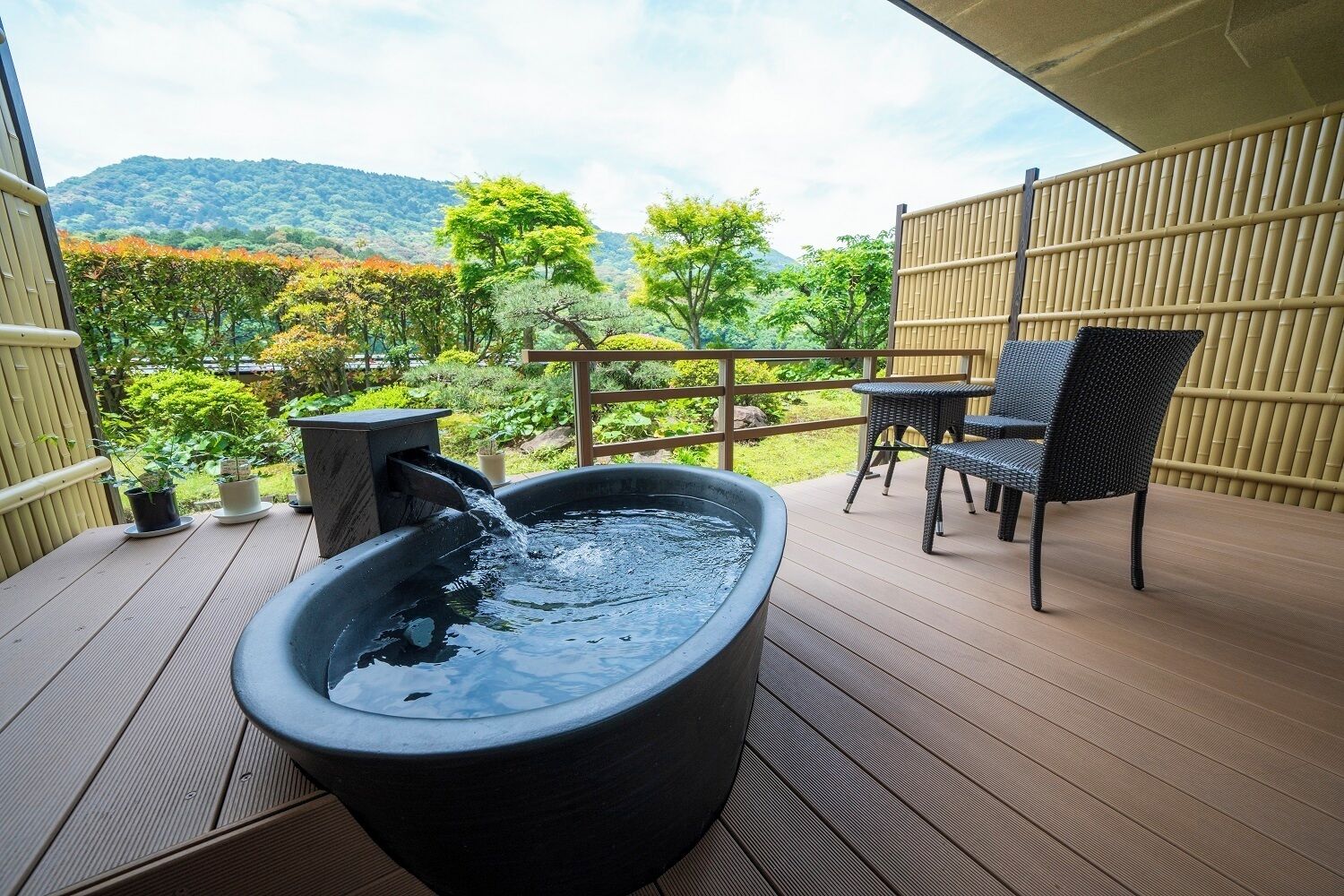 Un jacuzzi en una terraza de madera con valla de bambú y sillas, con vistas a un jardín exuberante y colinas.
