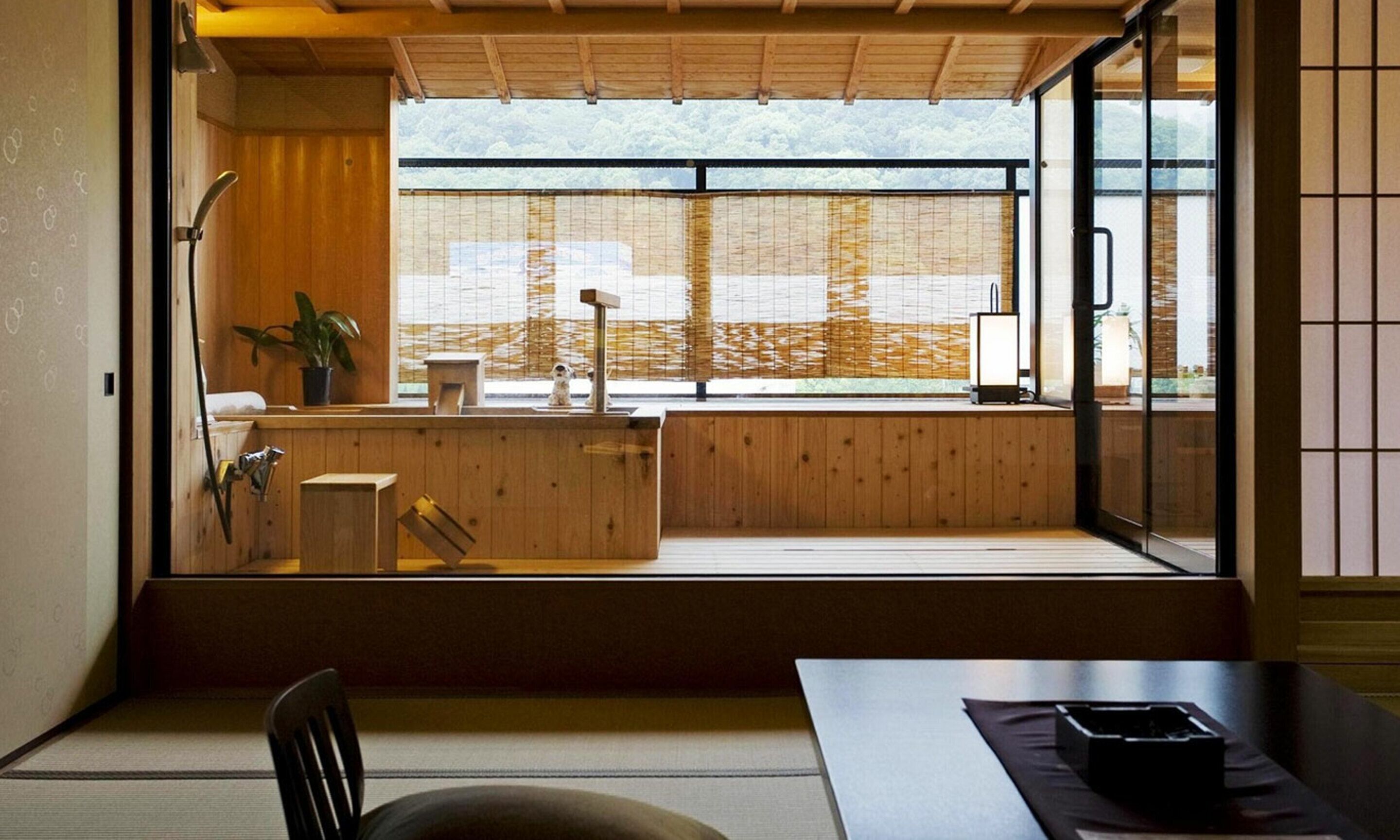 A traditional Japanese room with tatami flooring, a low wooden desk, a chair, a sliding shoji screen, and a view of a forested area.