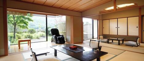 Ein traditioneller japanischer Raum mit Tatami-Böden, einem Tisch aus Tatami-Matten, einem schwarzen Massage-Sessel, einer Couch und Schiebetüren zu einem Garten.