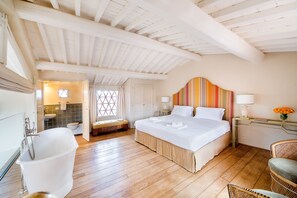 A bedroom with a large bed, a wooden floor, a white bathtub, and a window with curtains. - Borgo Del Cabreo (Greve in Chianti)