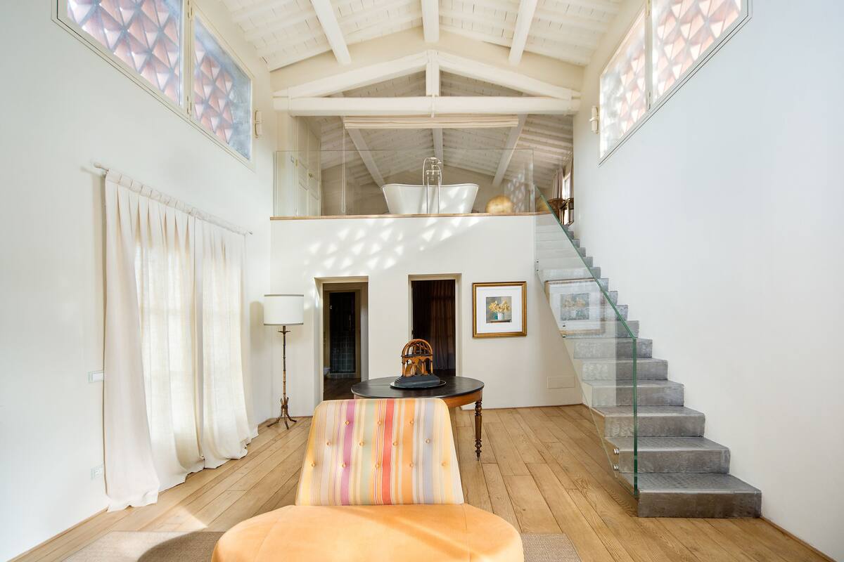 Una habitación amplia con piso de madera, una silla a rayas, una mesa con un adorno y una escalera con barandas de vidrio.
