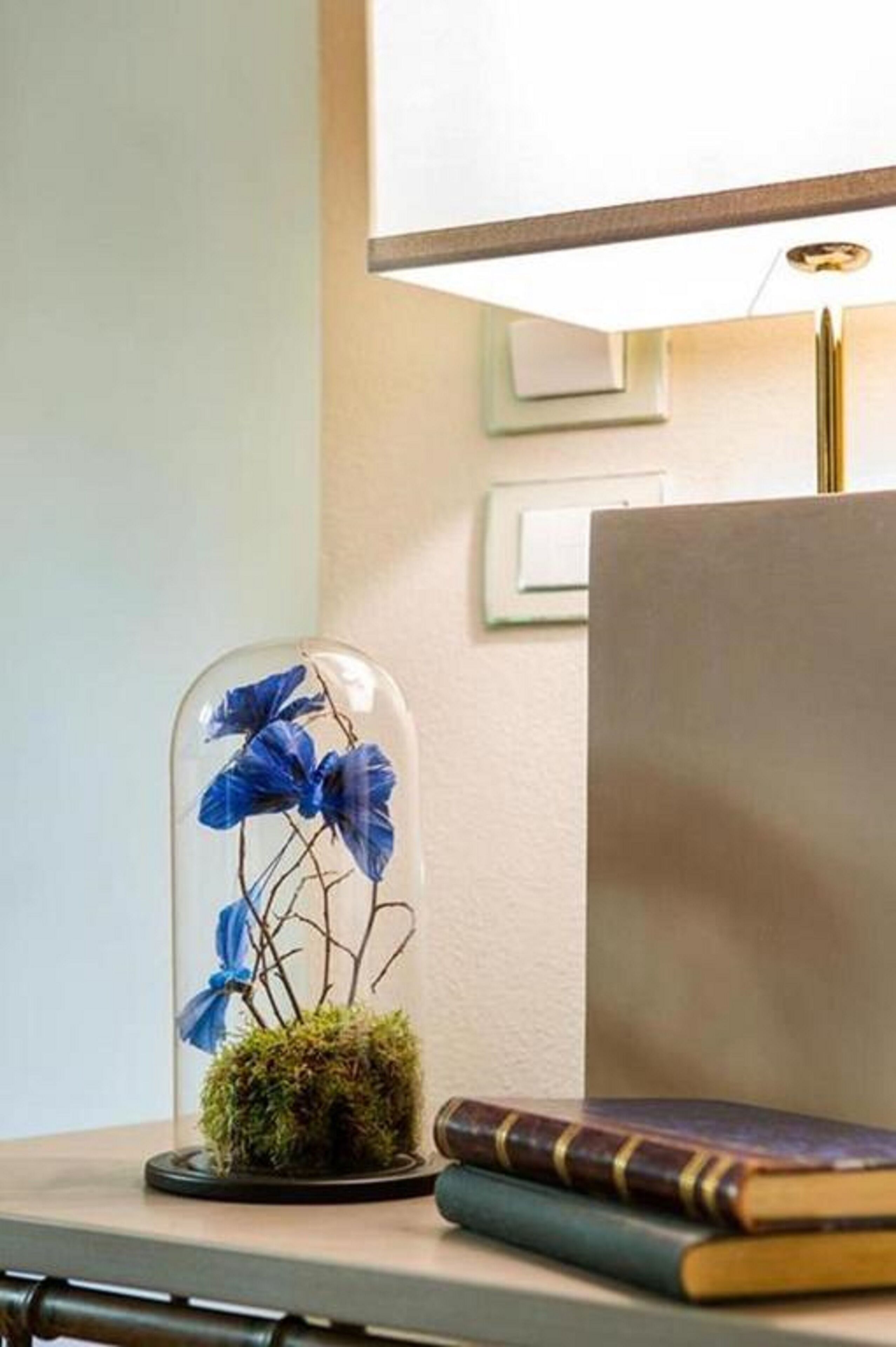 Ein Schreibtisch mit einer Glaskuppel, in der blaue Blumen und ein Moosfuß stehen, neben einer Lampe und einem Buch.