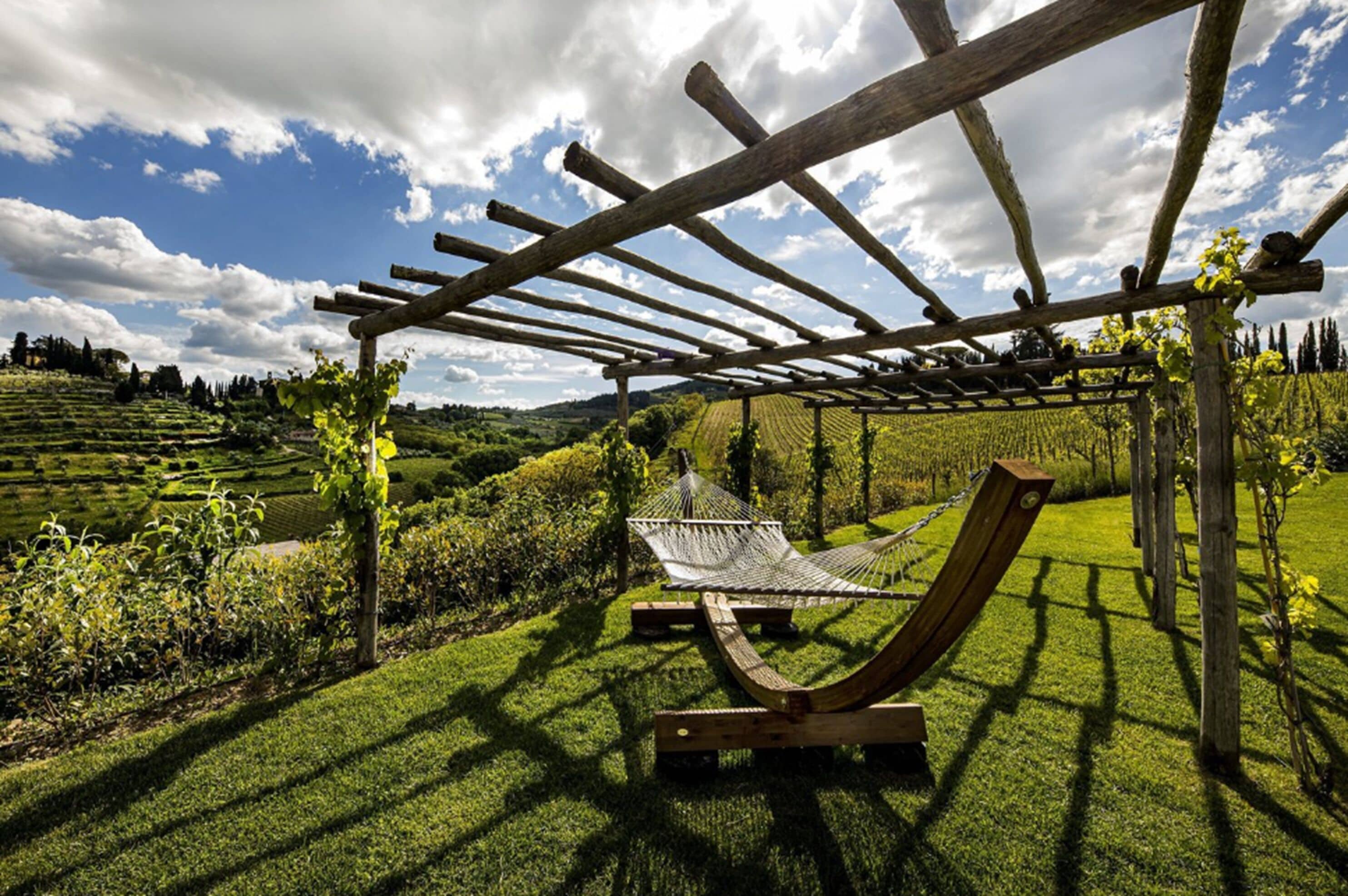 A hammock under a wooden pergola in a lush garden with a vineyard in the background.