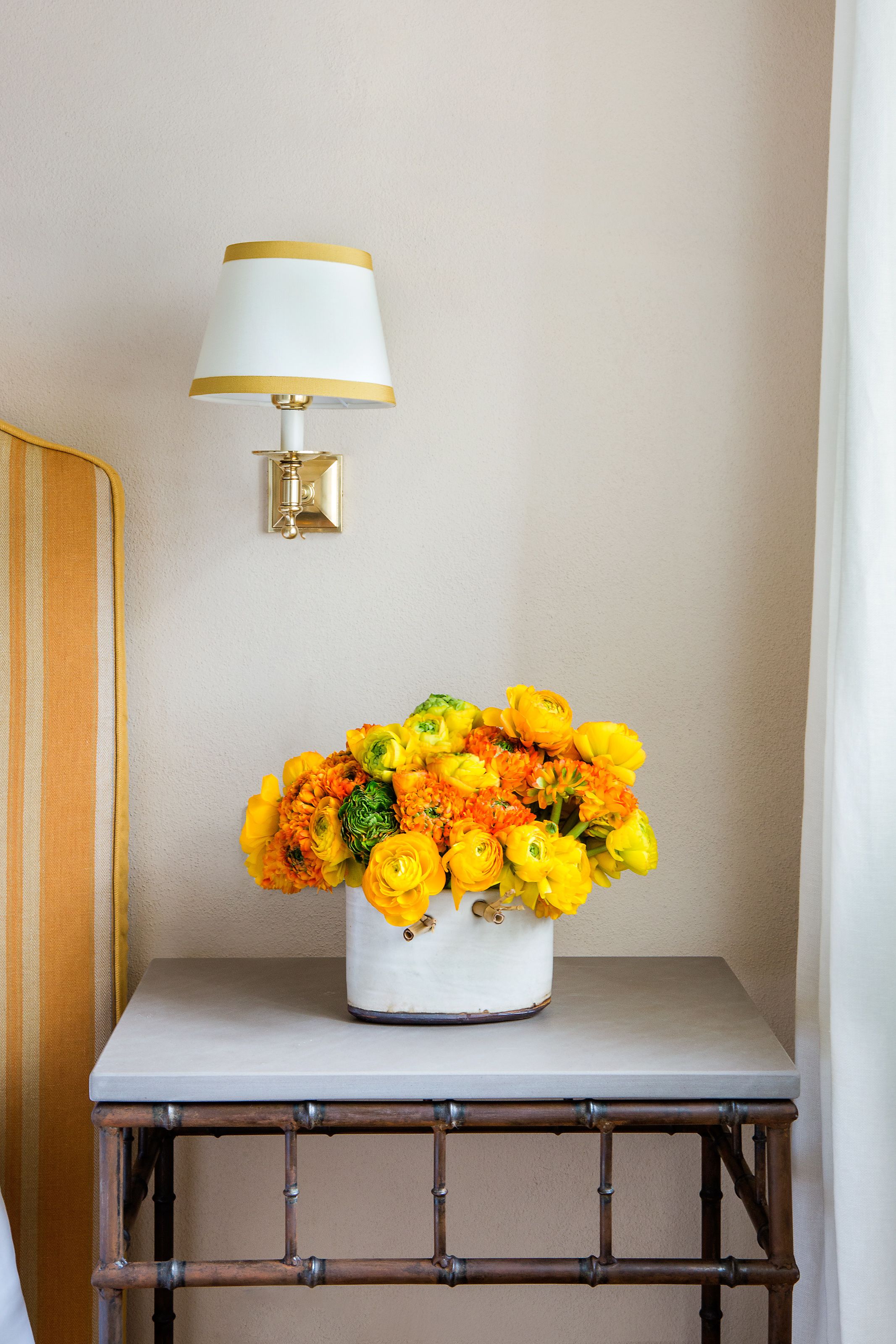Un vase blanc orné de fleurs jaunes, posé sur une petite table, une lampe fixée au mur et une chaise en bois.