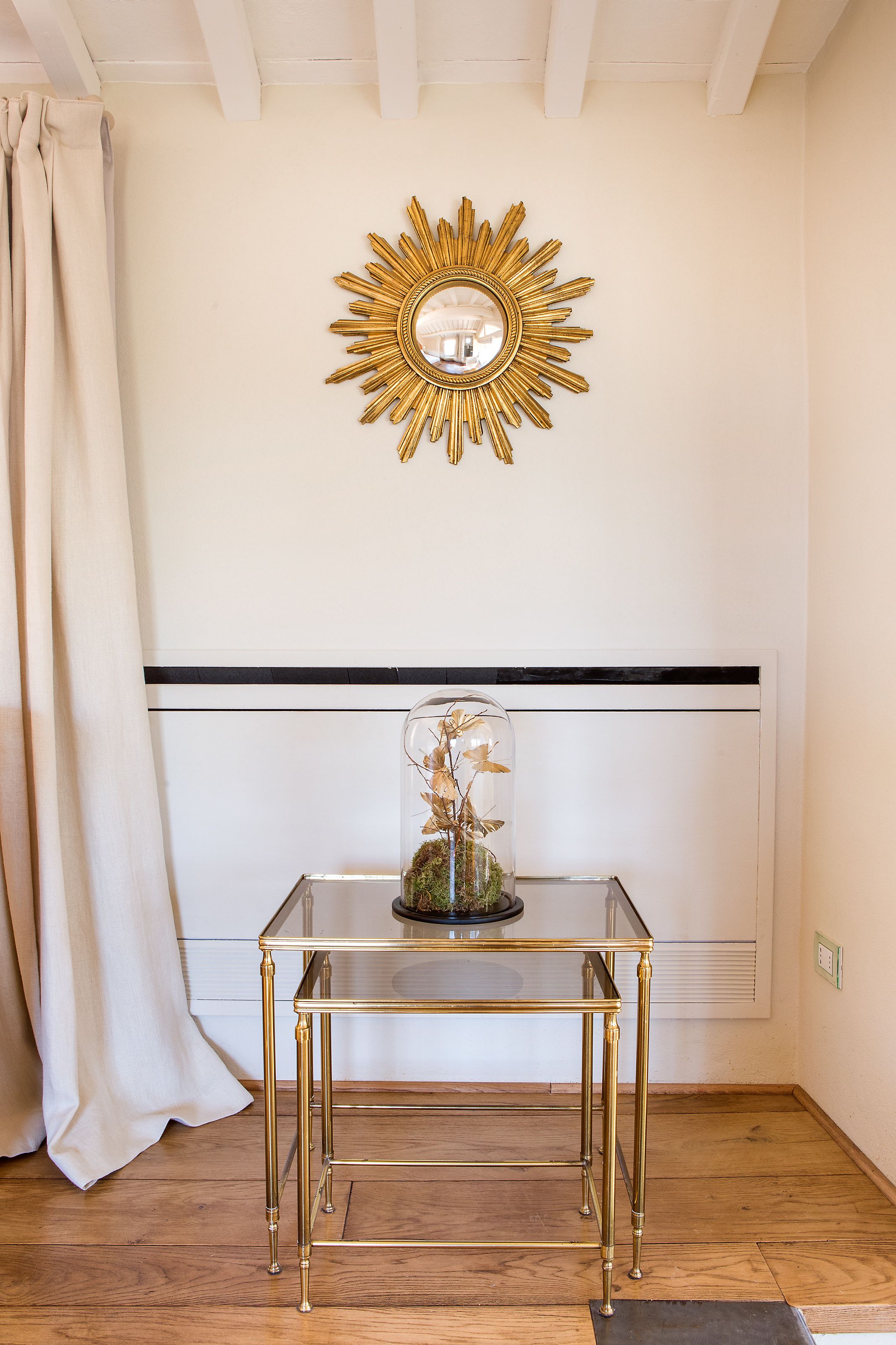 Una habitación con una mesa de vidrio, un espejo con forma de sol dorado y una cúpula de vidrio con flores secas.