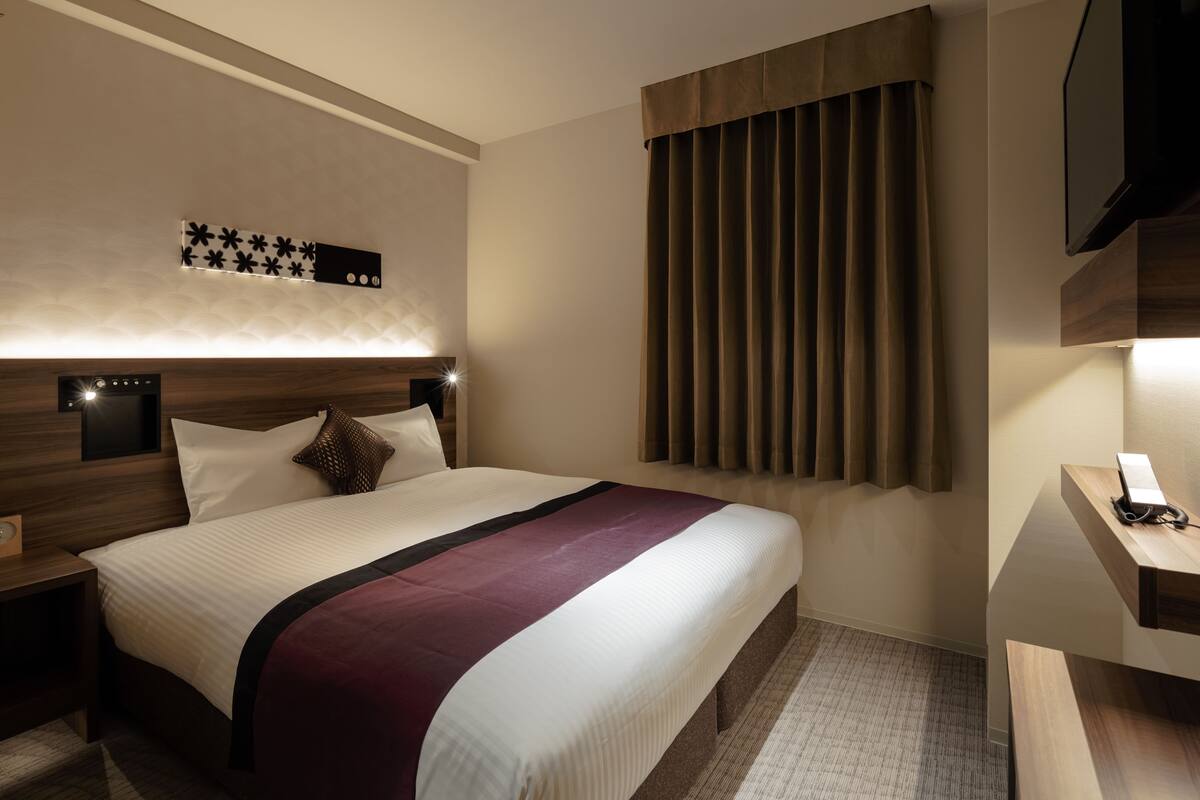 Habitación de hotel con una cama grande, una mesita de noche, un televisor de pantalla plana y un ventanal con cortinas.