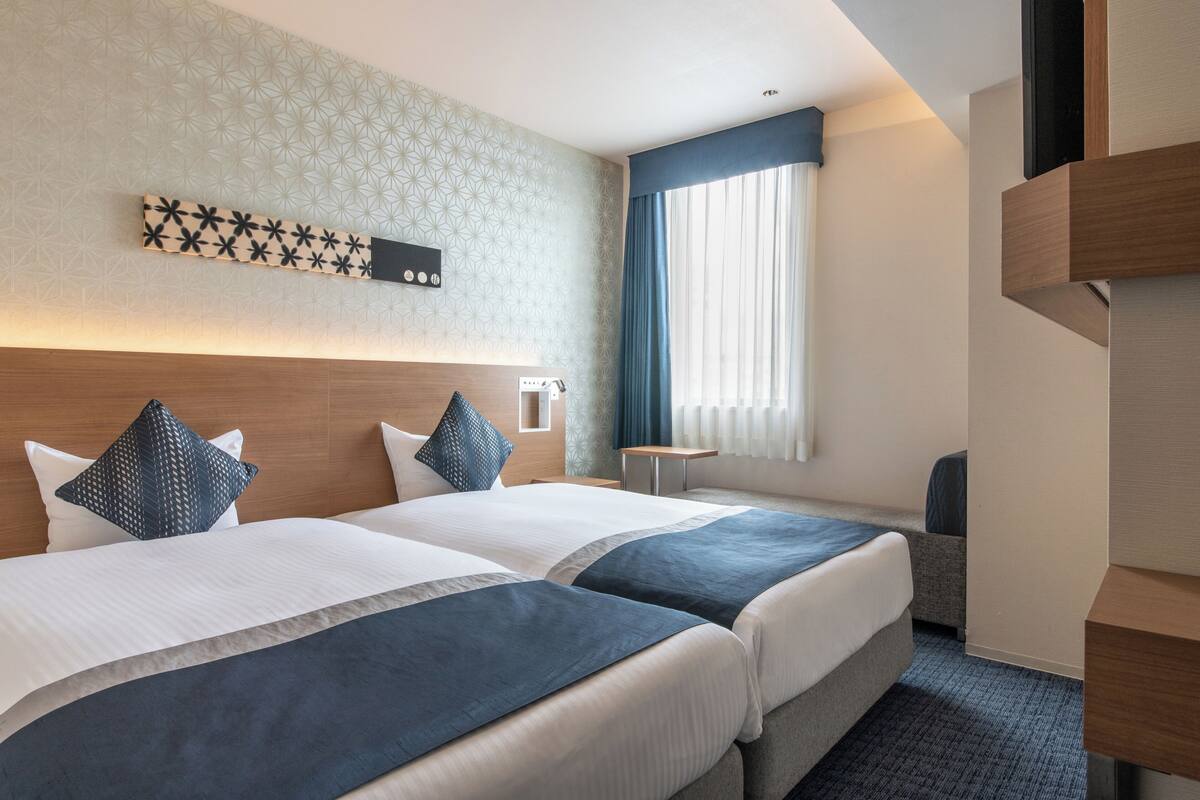 Habitación de hotel con dos camas, una mesita, una silla, una ventana con cortinas y una lámpara fijada a la pared.
