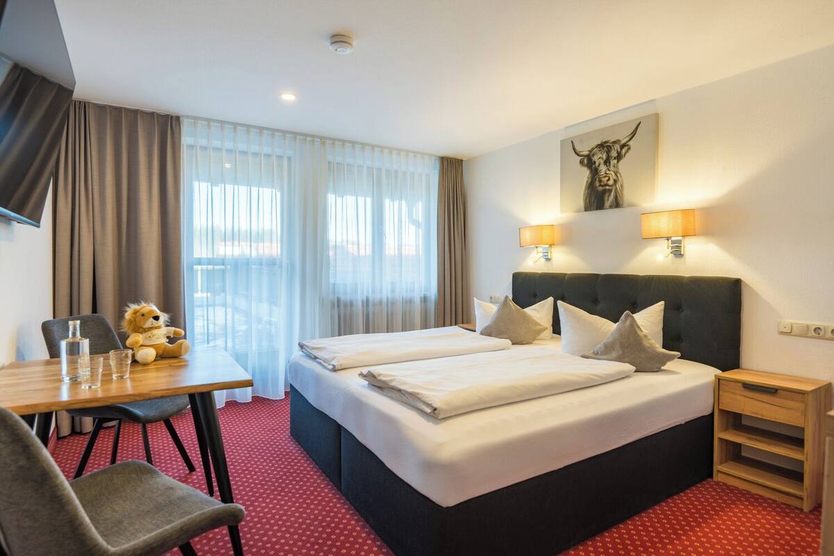 Une chambre d’hôtel avec un grand lit, deux lampes de chevet, un bureau avec une chaise, un ours en peluche et un tableau au mur.