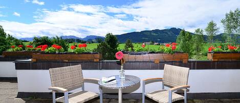 A terrace with two wicker chairs and a small round table, overlooking a garden with red flowers and a mountainous landscape.