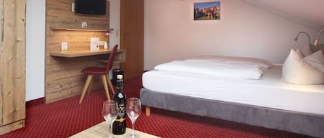 A hotel room with a bed, a wooden desk with a magazine, a bottle of wine, and a TV on the wall.