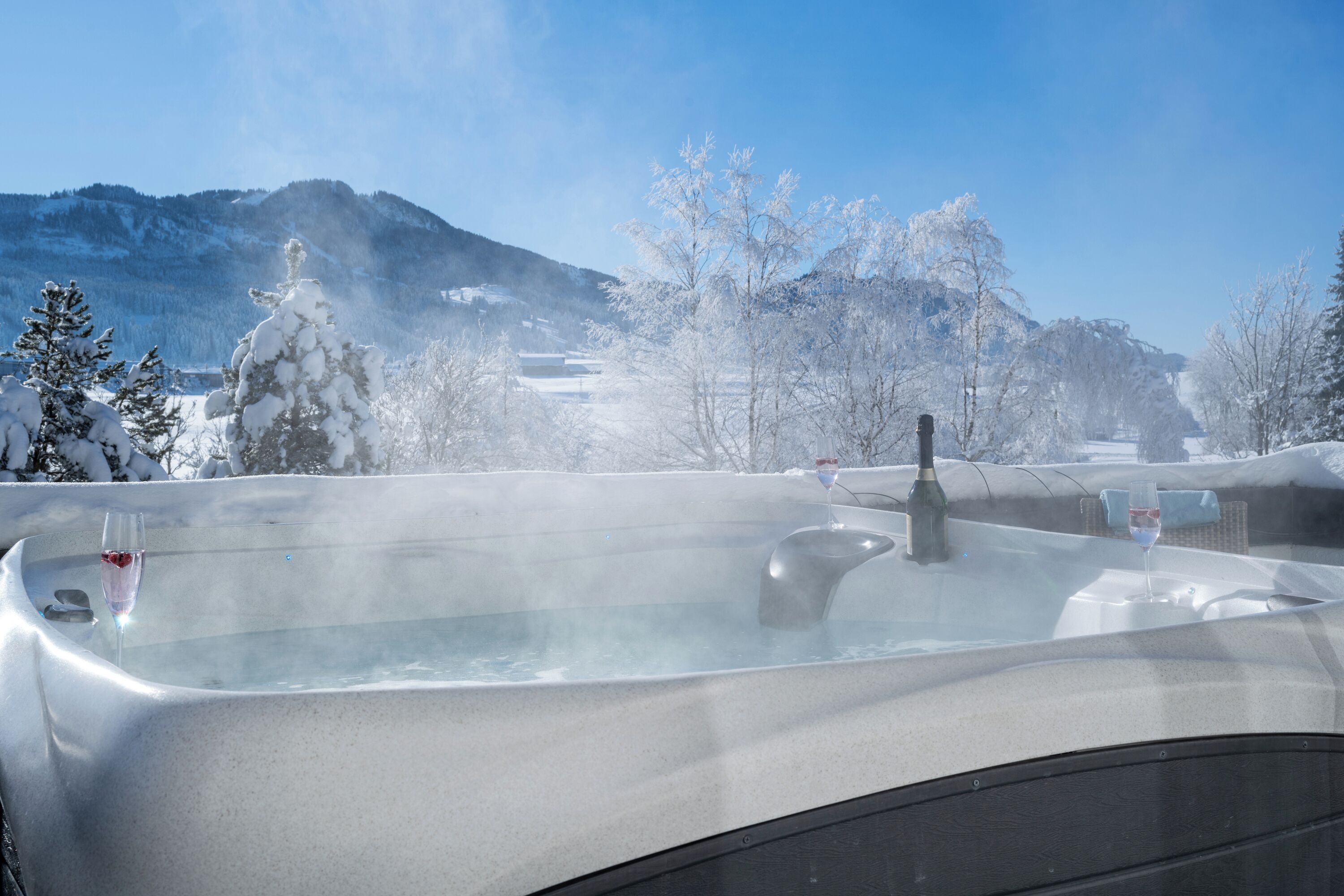 A hot tub with a glass of wine, surrounded by snow-covered trees and mountains.