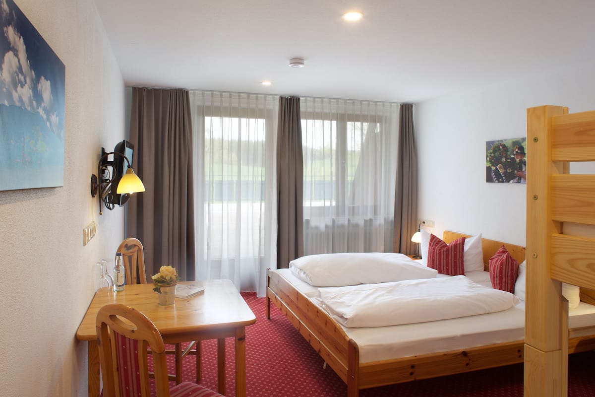 Une chambre d’hôtel avec un lit en bois, un bureau avec une chaise, une table ornée d’un vase de fleurs et un tableau accroché au mur.