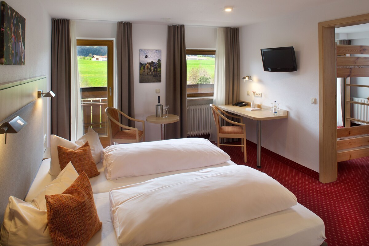 Een hotelkamer met twee bedden, een bureau, een stoel, een televisie en uitzicht op een groen landschap.