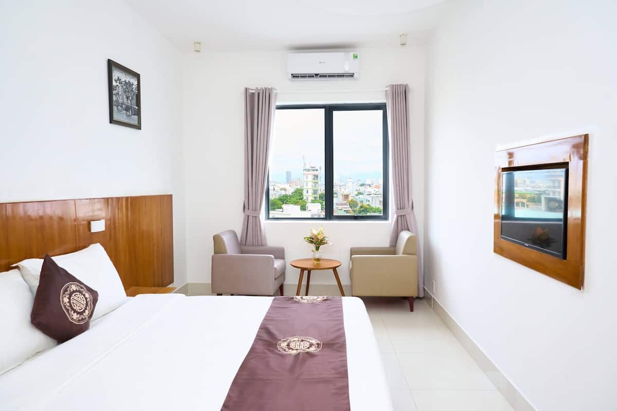 Quarto de hotel com cama, duas poltronas, uma mesinha com um vaso, janela com cortinas e TV fixada na parede.