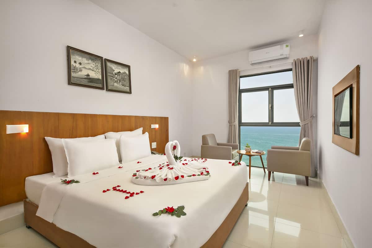 Quarto de hotel com uma cama, uma cadeira, uma mesinha e uma janela grande com vista para o mar.