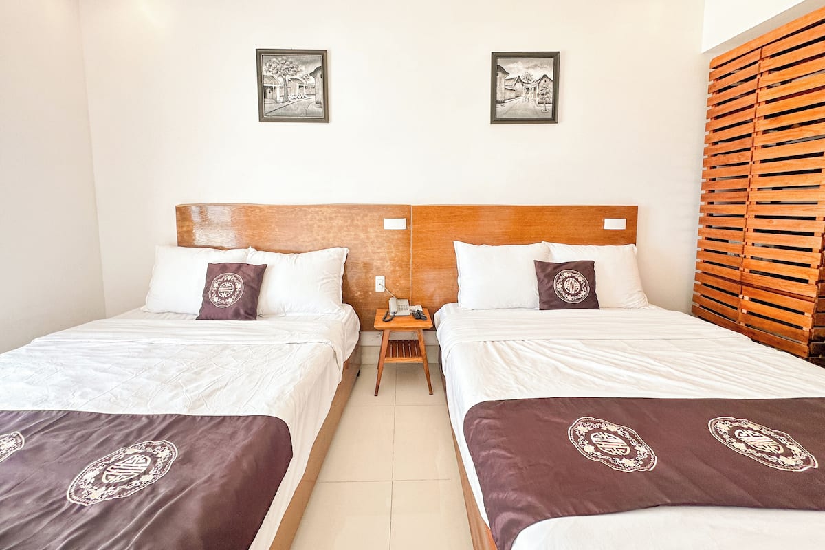 Quarto de hotel com duas camas, cada uma com cabeceira de madeira, roupa de cama branca e almofadas decorativas. Há duas imagens emolduradas na parede acima das camas.
