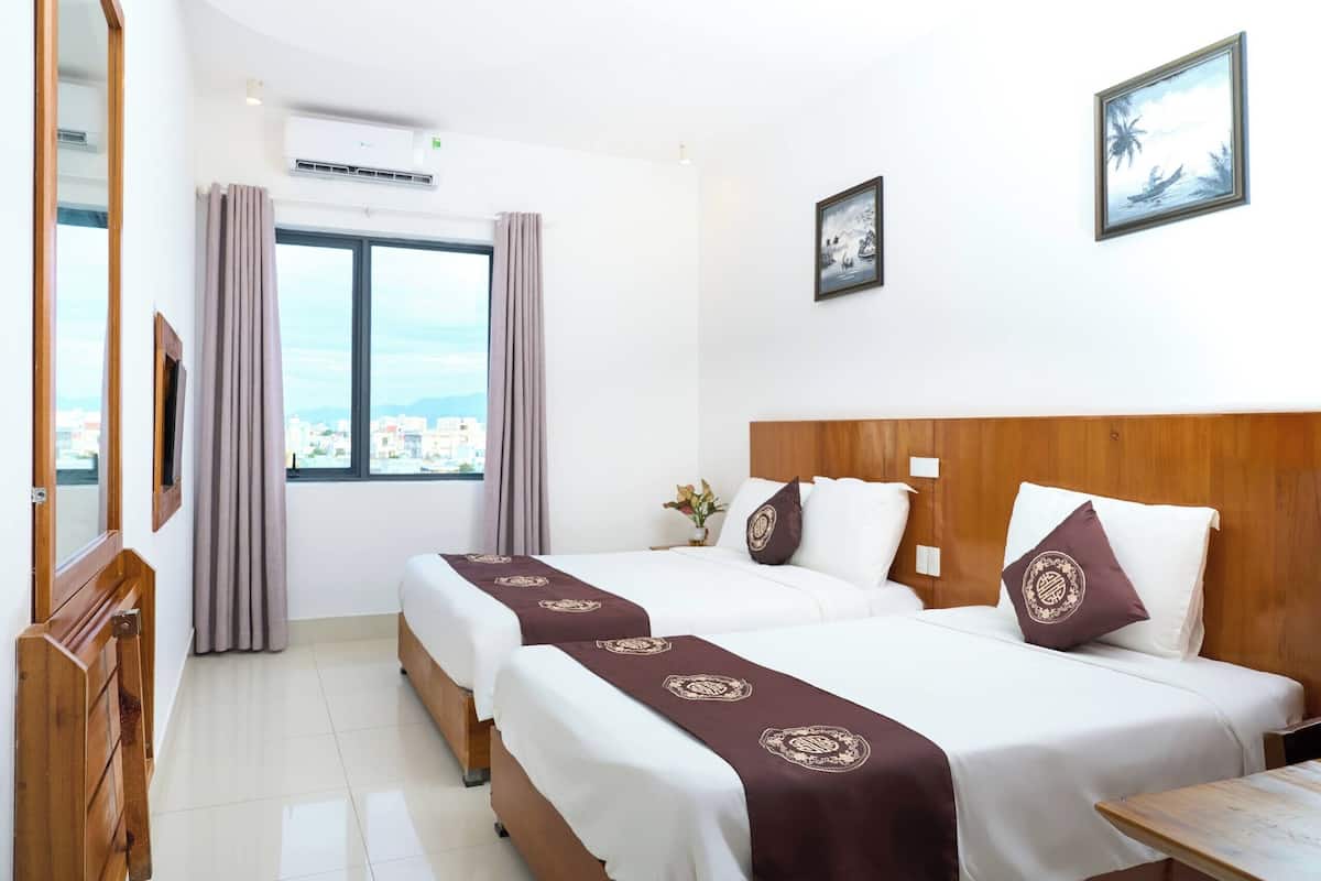 Quarto de hotel com duas camas, cabeceira de madeira, uma janela grande com cortinas e vista para a cidade.