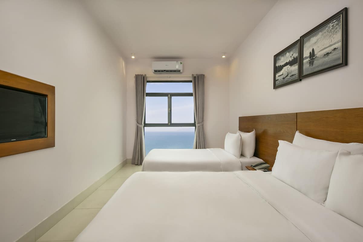 Quarto de hotel com duas camas, uma janela grande com cortinas, um aparelho de ar condicionado e duas imagens emolduradas na parede.